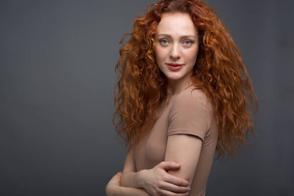 Счастливая кудрявая рыжая женщина с красивой улыбкой, держащая своими руками волосы. На изолированном сером фоне. Концепция: счастье и эмоции

