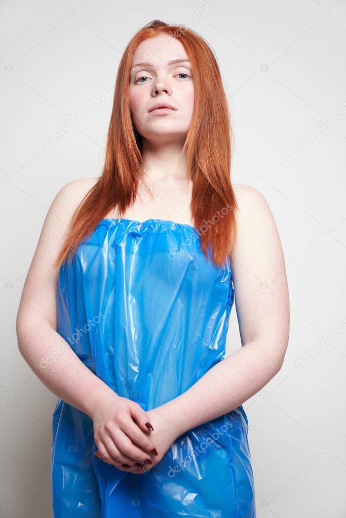Portrait of a young red-haired woman on a gray background. In a blue plastic bag. The concept of plastic pollution of nature. Excess plastic in a person's life                                       