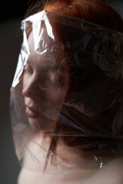 Kafasında plastik torbayla gri arka planda genç bir kızıl kızın portresi. Doğanın plastik kirliliği kavramı. Bir insanın hayatında aşırıya kaçan plastik                                        
