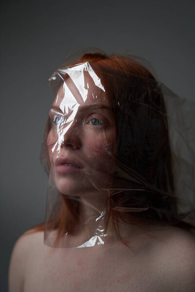  Portrait of a young redhead girl on grey background with a plastic bag on her head. The concept of plastic pollution of nature. Excess plastic in a person's life                                       