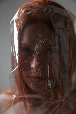 Kafasında plastik torbayla gri arka planda genç bir kızıl kızın portresi. Doğanın plastik kirliliği kavramı. Bir insanın hayatında aşırıya kaçan plastik                                        