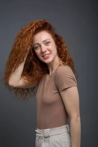 Happy curly red-haired woman with beautiful smile, holding her hair with her hands. On an isolated gray background. Concept: happiness and emotions