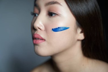 Profile of a young beautiful Asian girl with a cheek smeared with blue oil paint in the form of a horizontal stripe. Close-up photo. On a gray background, a place to copy                              