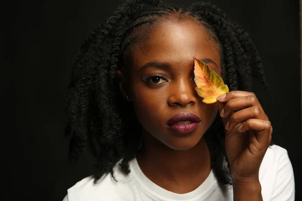 Portrait of a young beautiful African woman, with thick black curly hair, on an isolated black background. A woman covers one eye with a yellow autumn leaf.