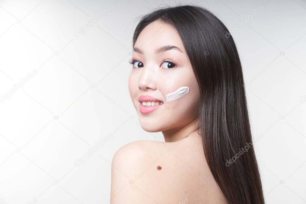  Portrait of a young Asian girl with a horizontal stripe on her cheek, smeared with white cream. Looks directly at the camera. On white background. The concept of skin care and beauty                                  