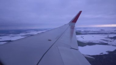 Pencereden uçağın kanadını görebilirsiniz. Arka planda bulutlar var. Uçak alçaktan uçuyor.