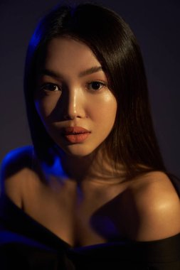 Portrait of a young beautiful Asian girl looking directly at the camera. The woman's hair and shoulders in colorful bright UV blue lights. On black background.                                  