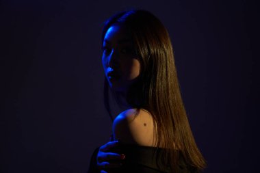  Portrait of a young beautiful Asian girl looking at the camera. A woman's face and shoulders in colorful bright UV-blue lights. On black backgrounds.                               