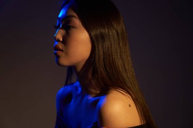  Profile of a young beautiful Asian girl, looking away. A woman's face in colorful bright UV-blue lights. On a gray background.                                 