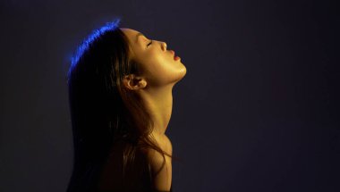  Profile of a young beautiful Asian girl looking up. Woman's hair in colorful bright UV blue lights. On black background.                                  