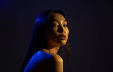 Profile of a young beautiful Asian girl, she looks directly at the camera. The hair and the back of the woman in bright , bright blue ultraviolet lights. On black background.                               