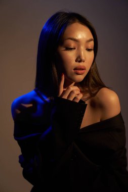 Profile of a young beautiful Asian girl, she looks away. The woman's hair and shoulders in bright bright ultraviolet blue lights. The hand is near the chin. On black background.                               