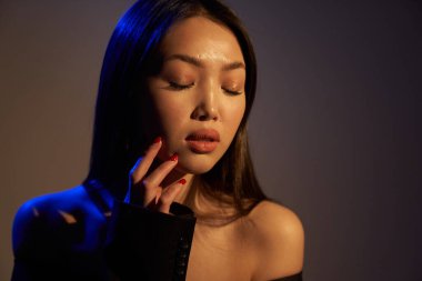 Profile of a young beautiful Asian girl, with her eyes closed. The woman's hair and shoulders in bright bright ultraviolet blue lights. The hand is near the chin. On black background.                               