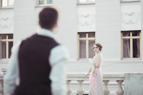 光の建物を背景にレトロなドレスを着た美しい女性が立っています 彼女は男を見て 彼が彼女に近づくのを待つ 歴史的復興 — ストック写真