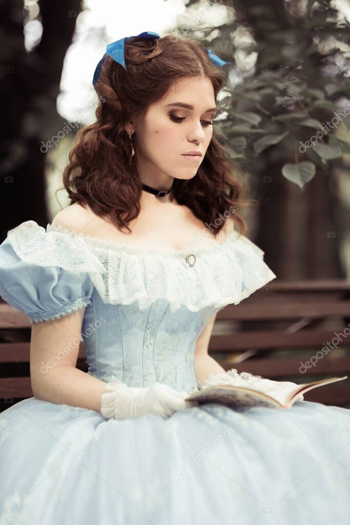 A young beautiful girl in a full blue dress, with curly hair, is sitting on a bench, holding a book in her hands and reading it. Against a tree. Historical reconstruction