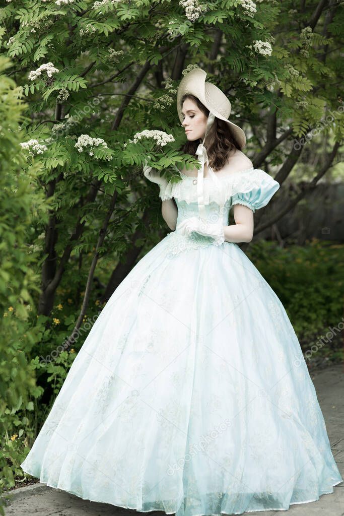 A beautiful girl in a puffy blue dress and hat, holding a lace umbrella in a folded state, looks away. Full-length photo. Historical reconstruction