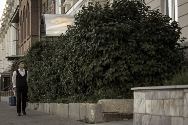 Genç bir adam bir binanın arka planında duruyor ve büyük yeşil çalılar. Sevgilisini bekliyor. Tarihi yeniden yapılandırma