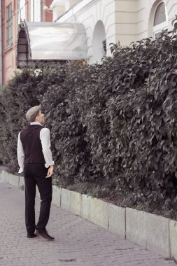 Genç bir adam bir binanın arka planında duruyor ve büyük yeşil çalılar. Sevgilisini bekliyor. Tarihi yeniden yapılandırma