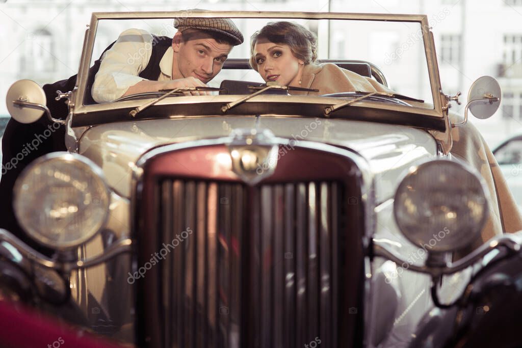 A young couple stands next to a retro car .They reach out to each other across the car and look out the windshield. Historical reconstruction