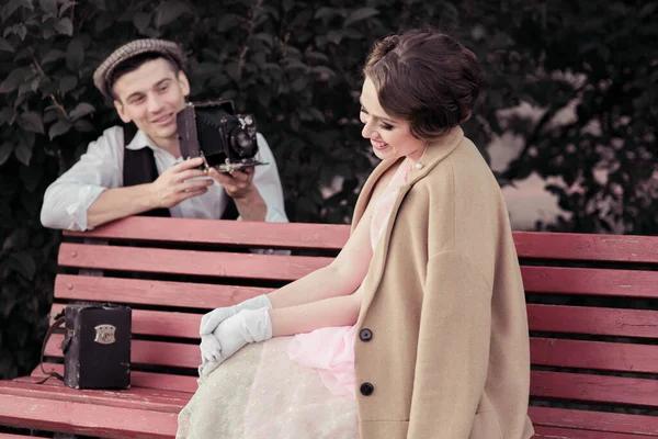 A young couple is photographed on a retro camera. A woman sits on a bench and looks away, a man photographs a stylishly dressed woman in retro style and looks at the woman.Historical reconstruction