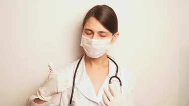Beyaz, tek kullanımlık koruyucu maskeli ve tek kullanımlık eldivenli bir kadın doktor önce sağ eliyle, sonra sol eliyle stetoskobu alıp başını sallıyor.
