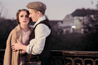 Genç bir çift bir çite, yeşil ağaçlara ve evlere karşı duruyor. Adam, kadına bakıyor. Tarihi yeniden yapılandırma. Retro kostümler.