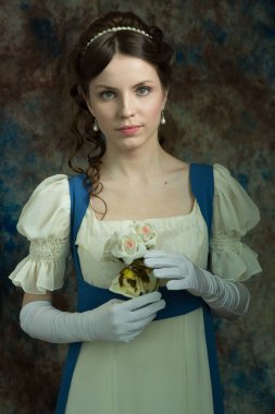 Beyaz ve mavi renkli uzun elbiseli genç bir kız. Doğrudan kameraya bakıyor. Beyaz çiçekler tutuyordu. Eski moda elbiseli bir kadın. Tarihi yeniden yapılandırma