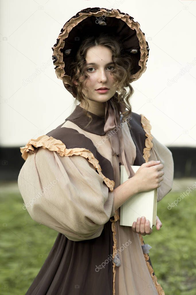 A young girl in a hat stands against the background of the forest. Holding a book, looking directly at the camera, in a retro dress. Historical reconstruction