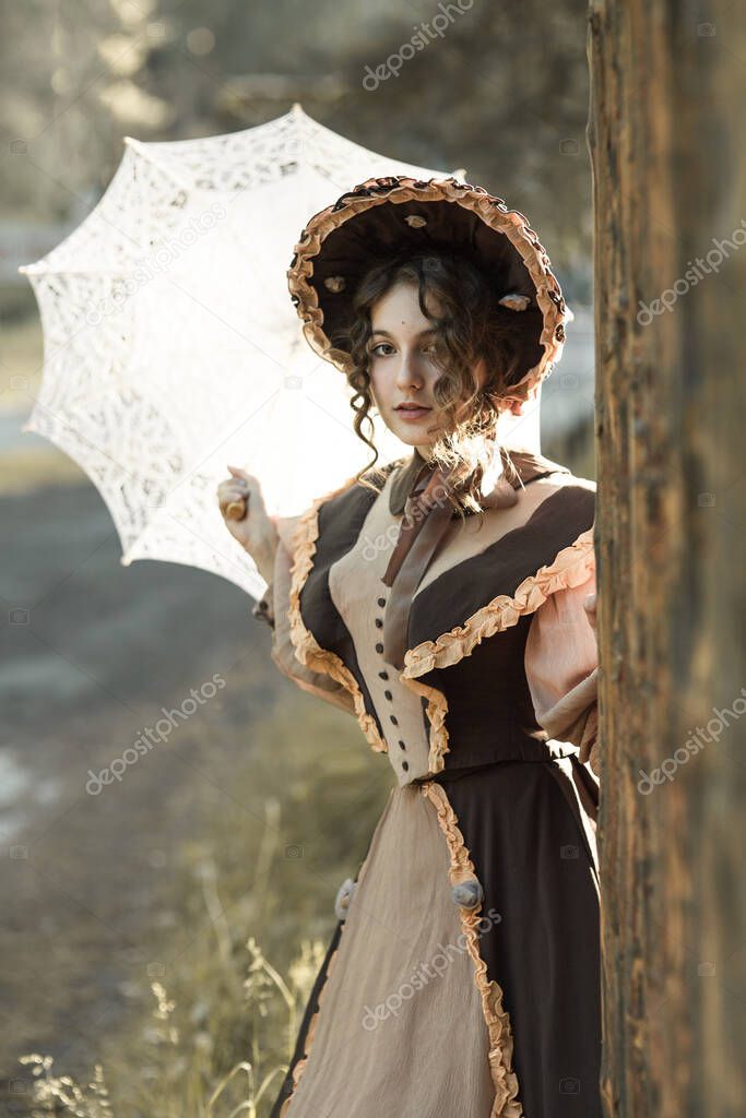 A young girl in a hat, standing behind a tree and looking directly at the camera, in a retro dress. Behind her is a white lace parasol. Historical reconstruction