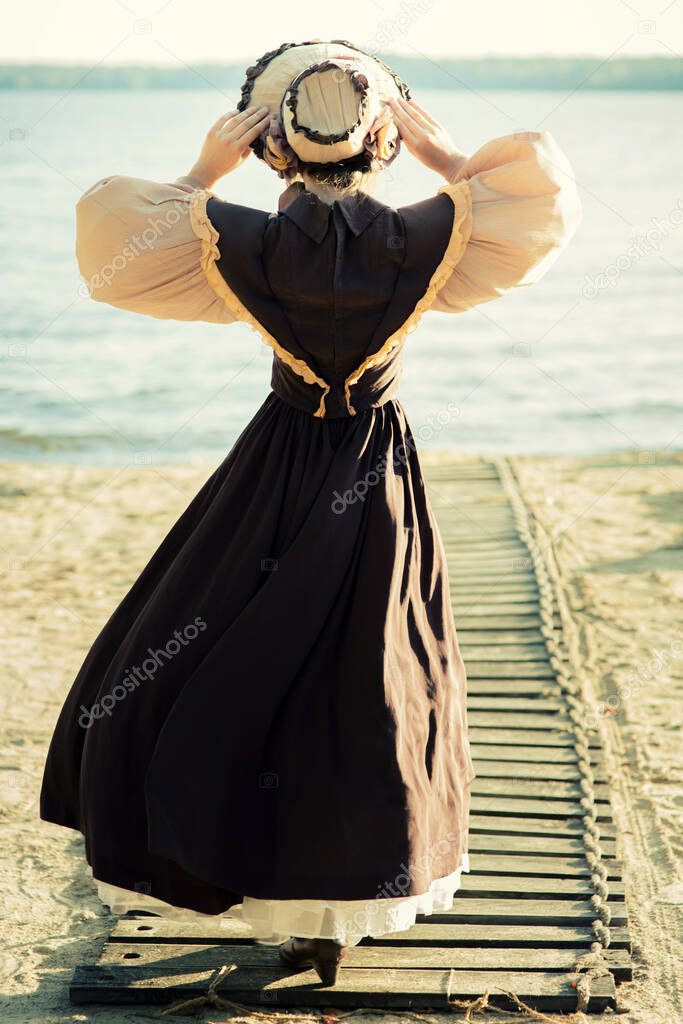 A young girl in a hat, standing full-length with her back to the camera, in a retro dress, holding her hat with her hands. Historical reconstruction