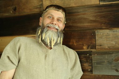 Sakallı, orta yaşlı, gri keten yelekli bir adamın stüdyo portresi, gülümseyen, başka tarafa bakan, sakalında mısır kulakları, kafasında dekoratif ip. Ekin konsepti