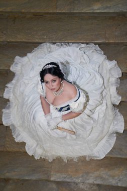  Güzel bir kız, romantik bir elbise içinde, eski moda. Koyu renk saçlı. Tam gelişim halinde, merdivenlerin arkasında oturuyor. Tarihi yeniden yapılandırma                                       