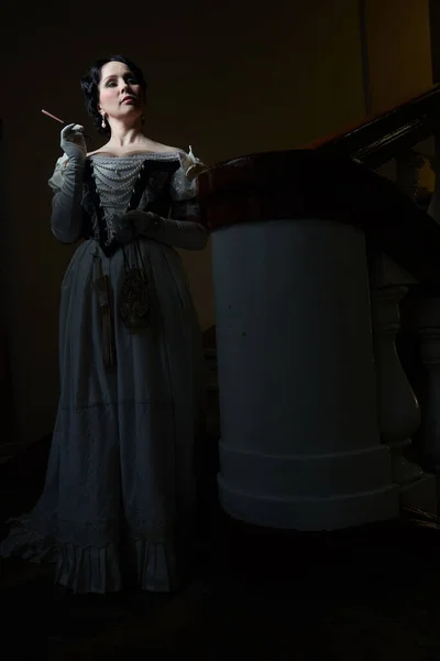 Beautiful girl, in a romantic dress, retro style.With dark hair. Stands against the background of the stairs in the theater, and looks away. Holding a mouthpiece and a purse. Historical reconstruction