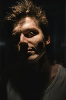 Beyaz tişörtlü bir adamın portresi, doğrudan kameraya bakıyor. Işık yüzün bir kısmına düşer.