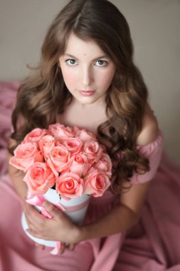 Pembe uzun elbiseli, uzun kıvırcık saçlı, pembe güllü genç bir kızın portresi, oturur ve doğrudan kameraya bakar.