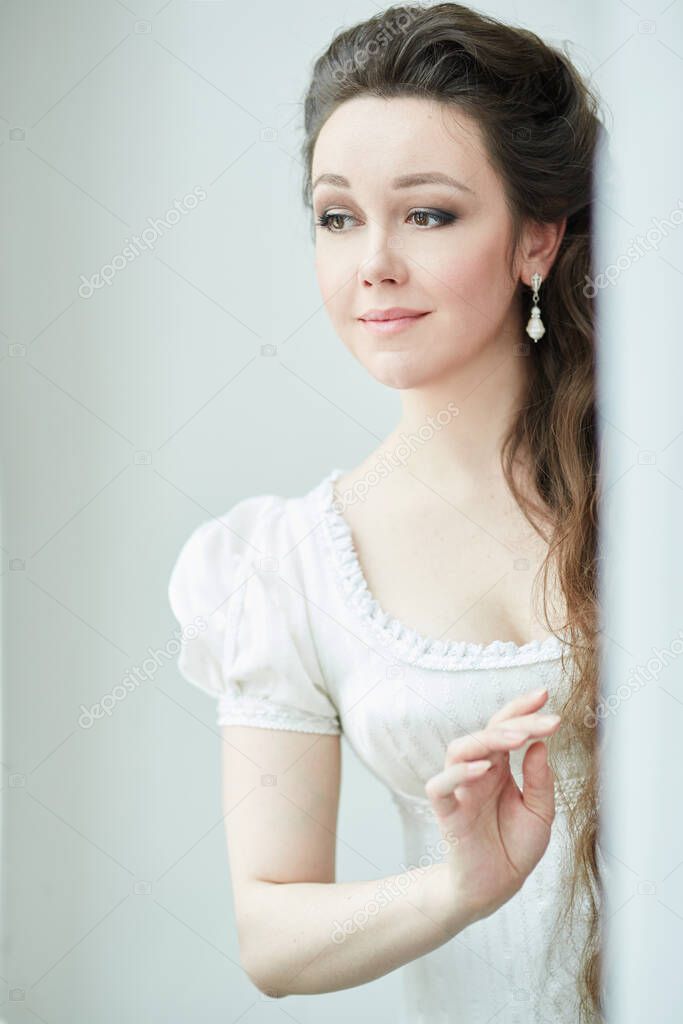 A young girl with long hair, in a white dress, stands outside the door and looks away, in a retro dress. Historical reconstruction