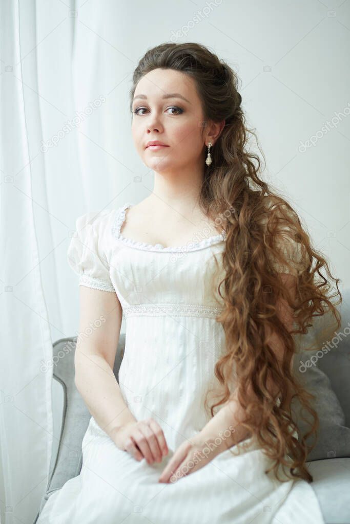 A young girl with long hair, in a white dress in empire style, sits and looks directly at the camera, in a retro dress. Against the background of white curtains. Historical reconstruction