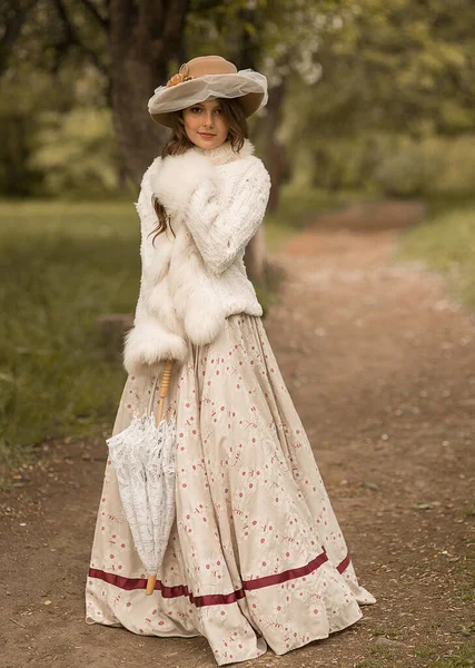 一个戴着帽子 头戴长发 身穿白色长裙 头戴花边雨伞的漂亮姑娘的画像 站在森林小径上 直接看着摄像机 历史重建 — 图库照片
