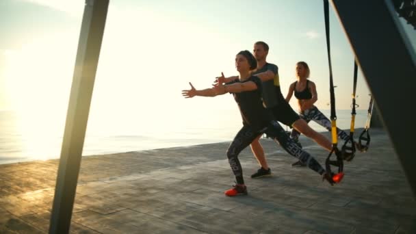Pessoas esportivas treinando agachamentos com trx perto do mar contra o sol. Movimento lento . — Vídeo de Stock