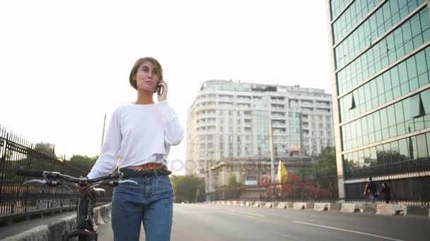 Joven mujer llevando su bicicleta habla por teléfono — Vídeos de Stock