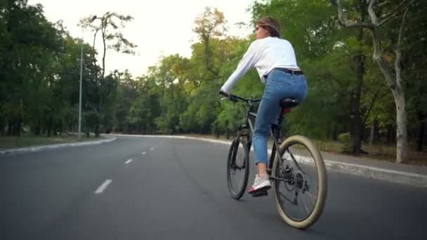 Bakifrån ung kvinna rider en cykel genom parken — Stockvideo