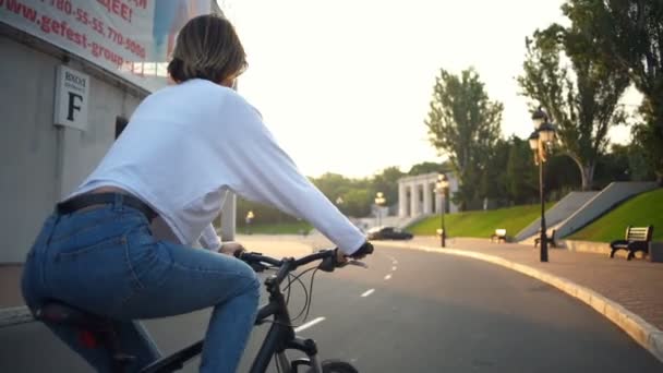 Back view young female riding a bike through the park in sunlight — Stock Video