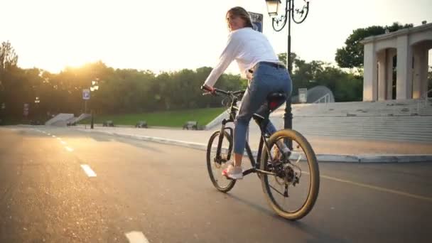Jovem fêmea monta uma bicicleta no parque sorrindo para a câmera às vezes — Vídeo de Stock