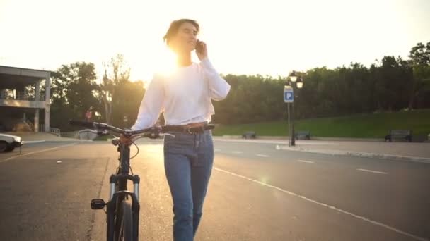 Jovem fêmea carregando sua bicicleta e falando na vista frontal do telefone — Vídeo de Stock