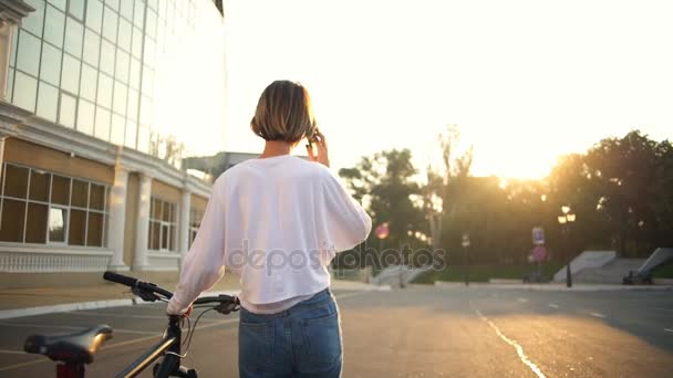 Joven hembra llevando su bicicleta y hablando en la vista posterior del teléfono — Vídeos de Stock