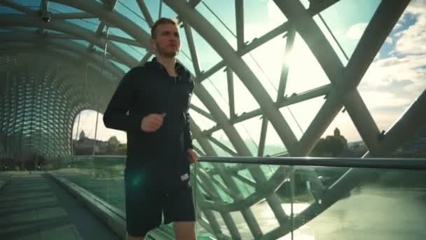 Jovem homem bonito com fones de ouvido correndo através da ponte moderna câmera lenta — Vídeo de Stock