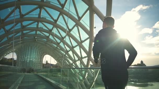 Jovem homem apto com fones de ouvido correndo através da ponte moderna câmera lenta — Vídeo de Stock