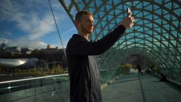 Jonge knappe man met hoofdtelefoon maken van een video-oproep op de brug slow motion — Stockvideo