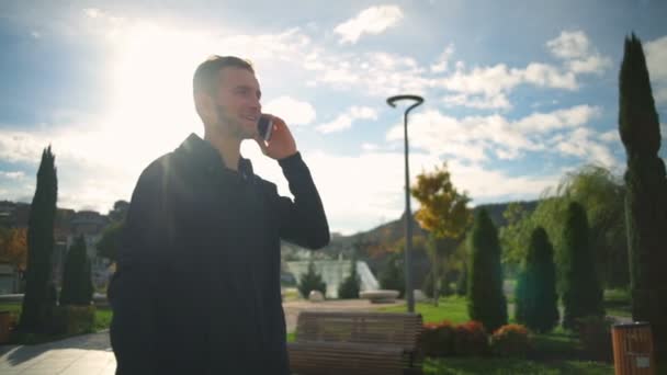 Um jovem caminhando em um belo parque e falando no telefone câmera lenta — Vídeo de Stock