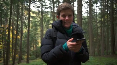 Onun elinde bir akıllı telefon ile ayakta ve bir ormanda gülümseyen bir kadın